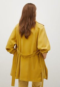 Veste jaune ceinturée à manches larges, confectionnée dans un tissu lisse, dotée d'une ceinture à la taille et d'un design épuré, vue de dos.