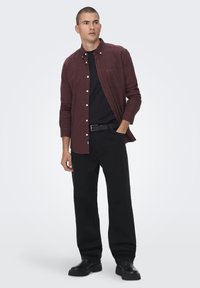 Chemise boutonnée bordeaux superposée sur un t-shirt noir, associée à un pantalon et des chaussures noires, avec une poche avant sur la chemise.