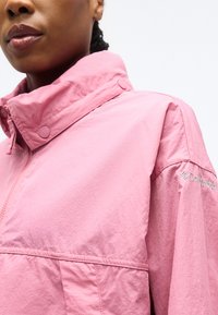 Γυναίκα με ροζ μπουφάν Columbia με ψηλό κολάρο και ορατό φερμουάρ και κουμπιά κούμπωμα στο κολάρο.