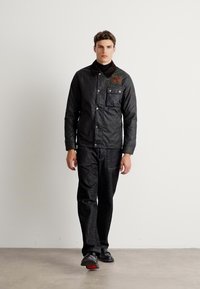 Veste matelassée noire avec un col montant, une poche avant et un écusson logo. Associée à un pantalon texturisé sombre et des chaussures noires.