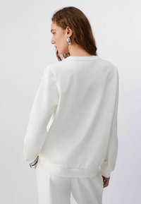 Femme aux longs cheveux bruns portant de grandes boucles d'oreilles argentées en forme de cercle, un sweat-shirt blanc à manches longues et un pantalon blanc, debout devant un fond uni.