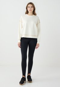 Felpa bianca con rifiniture dorate, scollo rotondo e spalle cadenti, abbinata a leggings neri e scarpe sportive nere.