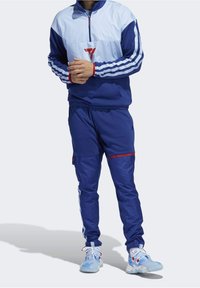 Survêtement athlétique bleu avec veste à col montant zippée au quart, accents rayés, et pantalon assorti avec poches et détails rouges.