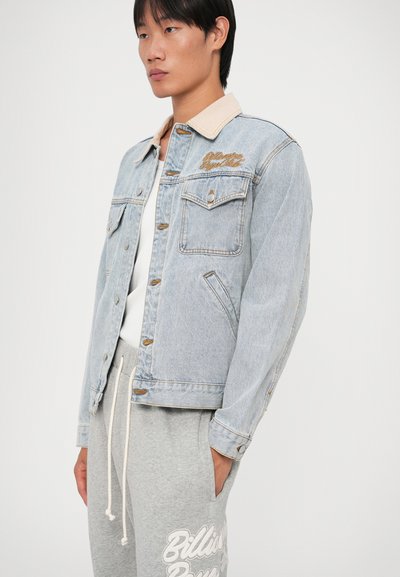 Lichtblauwe denimjack met een beige kraag, voorzien van twee borstzakken, oranje stiksels en geborduurde tekst op de borst.