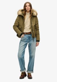 Olivowa puchowa kurtka z futrzanym kapturem, kremowy sweter w warkocze, jasnoniebieskie dżinsy o prostym kroju i brązowe półbuty na sznurówki.