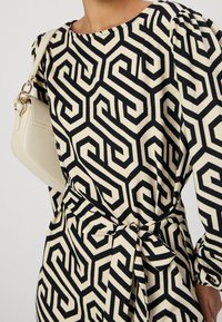 Schwarz-creamfarbene, geometrisch gemusterte Kleid mit puffärmel und passendem Gürtel, kombiniert mit einer kleinen creamfarbenen Handtasche über der Schulter.