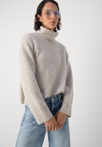 Lys beige ribbestrikket genser med høy hals og vide ermer, sammen med lyseblå høytlivs denimjeans. Glatt tekstur, avslappet passform.