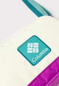Sac Columbia avec un corps crème, des accents violets et un patch logo turquoise. Il dispose d'un tissu texturé et d'une sangle ajustable teal.