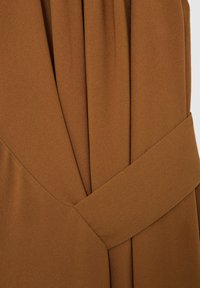 Brun stoffrakke med bredt bælte, der er snoet rundt om taljen, med detaljerede syninger og lagdelte folder i materialet.