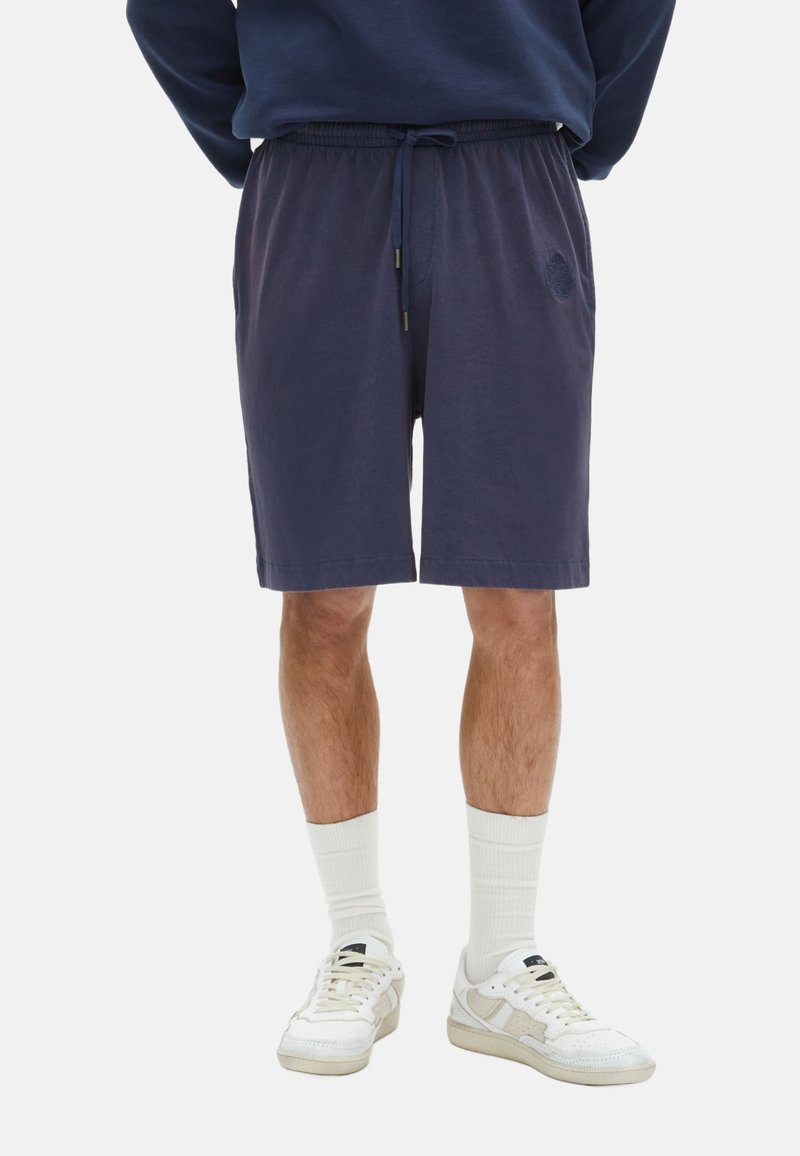 Marineblauwe katoenen shorts met elastische tailleband, koordsluiting, zijzakken en een logopatch. Gecombineerd met witte sokken en sneakers.