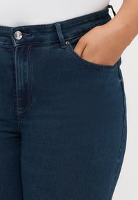 Dark blue high-waisted jeans with a smooth denim texture, featuring a front pocket and silver button closure detail.