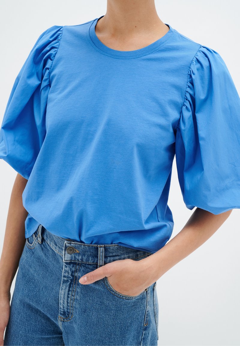 Blå bomullströja med puffärmar, rund halsringning och avslappnad passform, kombinerad med högmidjade denimjeans.