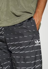 Svarta träningsshorts i slät tyg, med ett upprepat vitt "adidas"-logomönster och en vit trefoil-logotyp på sidan.