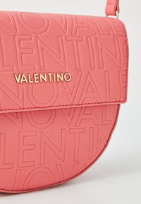 Rund rosa väska med präglad "VALENTINO"-logotyp. Texturerat lädermaterial med detaljer i guld. Kompakt och modern design.