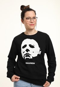 Felpa nera con un grande grafico bianco di un volto mascherato, con il testo "Halloween di John Carpenter" sottostante, realizzata in tessuto morbido.