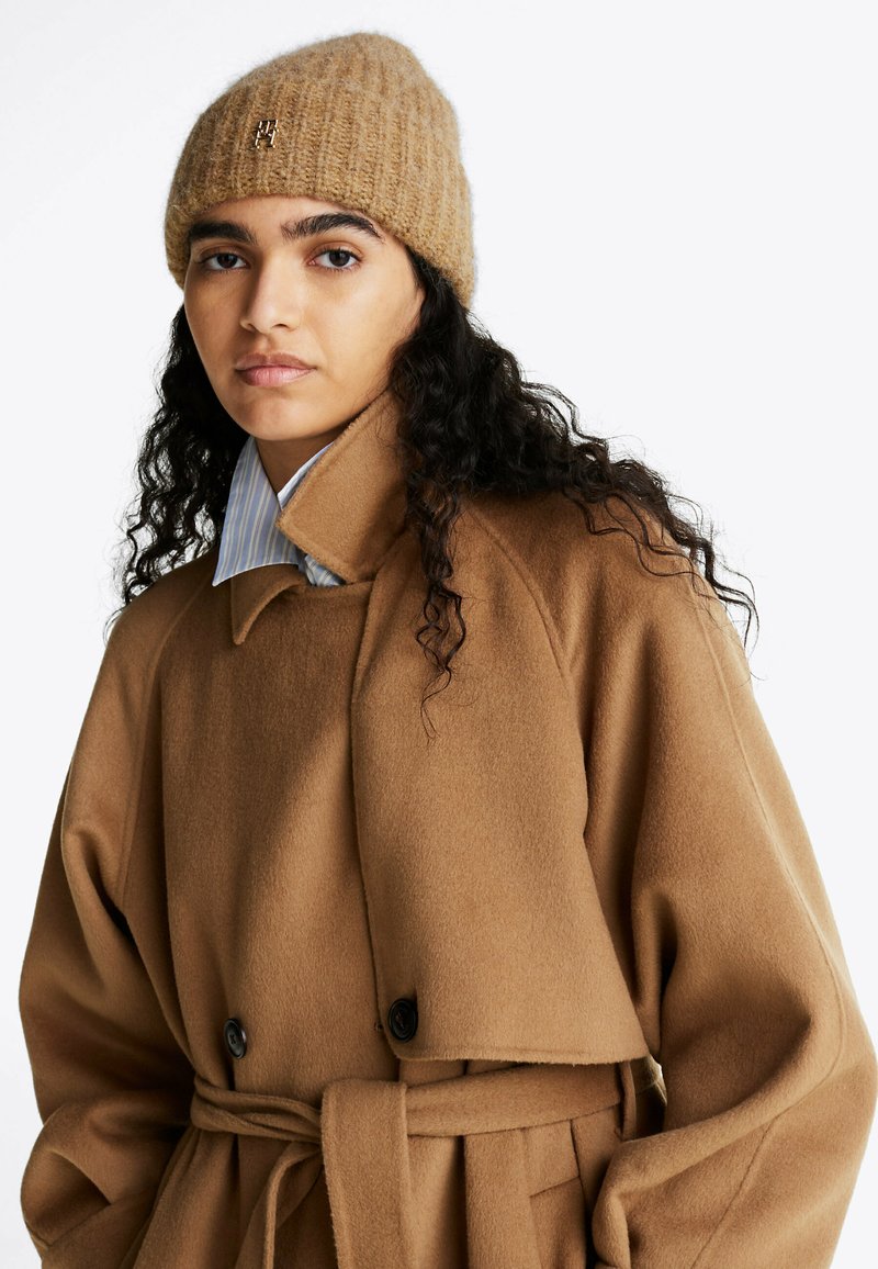 Bruine wolmix jas met een ceintuur in de taille, dubbele knopen en hoge kraag, gecombineerd met een tan gebreide beanie met een logodetail.