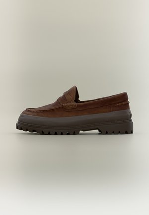 Mocassino in pelle marrone con suola spessa in gomma nera e suola esterna scolpita, mostrato di lato su uno sfondo neutro semplice.