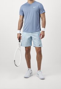 Miehen vaaleansininen Nike-urheilupaita ja kuvioidut sortsit. Pitelee tennismailaa, päällään valkoiset tenniskengät ja rannekkeet.