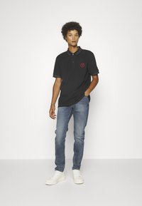 Camisa polo negra con un logo rojo, de mangas cortas y cuello, combinada con jeans de mezclilla azul claros y zapatillas blancas, sobre un fondo liso.