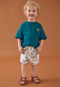 Chemise à manches courtes bleu sarcelle avec un motif de soleil, associée à un short crème orné d'imprimés colorés d'animaux et de palmiers, et des sandales marron.