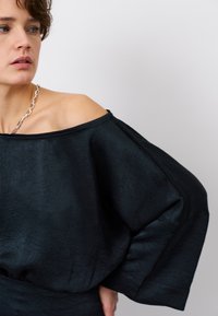 Zwarte off-the-shoulder top met een gestructureerd oppervlak, brede mouwen en een ontspannen pasvorm, gecombineerd met een zilveren kettinghalsband.