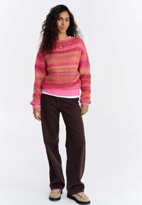 Kobieta o kręconych włosach, ubrana w różowo-pomarańczowy, paskowany sweter, ciemnobrązowe spodnie, beżowe buty i złoty naszyjnik, stojąca na białym tle.