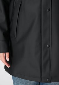 Zwarte waterdichte jas met een gladde textuur, drukknopen, zijzak en een losse pasvorm, aangevuld met blauwe denim die zichtbaar is aan de onderkant.