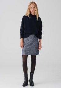 Donker marineblauwe trui met ribbelmanchetten, gecombineerd met een gestructureerde blauwe rok. Model draagt zwarte ondoorzichtige panty’s en enkellaarsjes met hak.