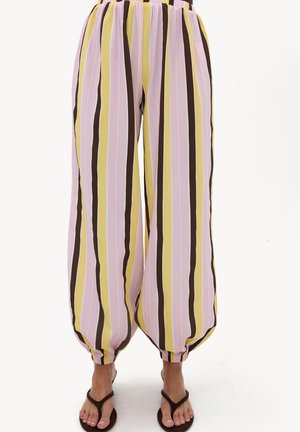 Pantalon large à rayures verticales rose, jaune, marron et blanc, resserré aux chevilles, porté avec des tongs marron foncé, montré de la taille vers le bas.
