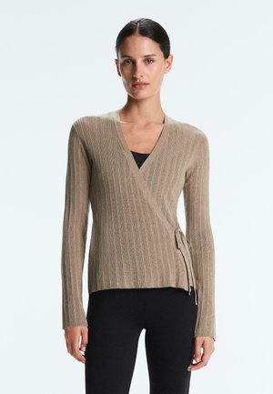 Žena má na sobě béžový žebrovaný zavinovací svetr s boční stuhou přes černý top a černé přiléhavé kalhoty, stojí na světlém pozadí.