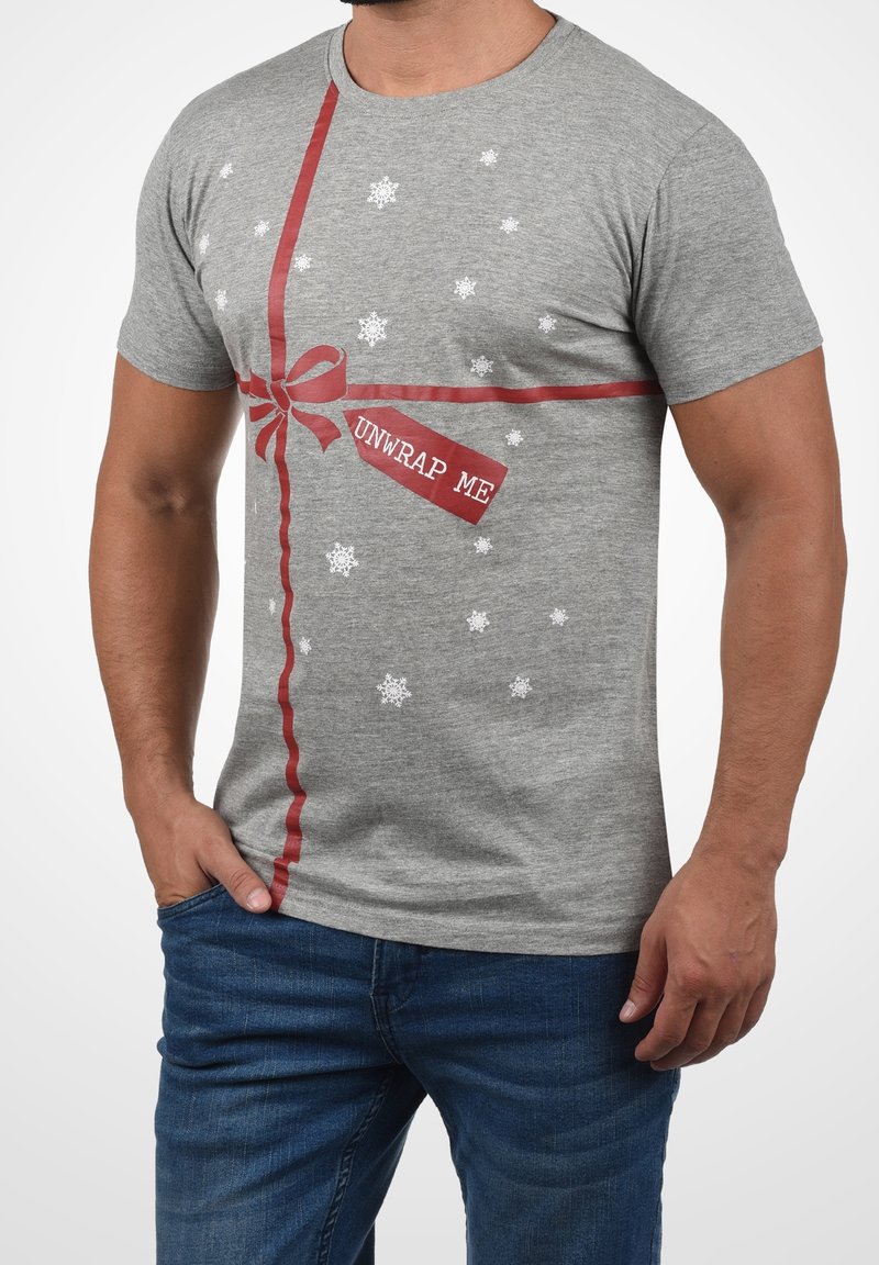Homme portant un t-shirt gris avec un imprimé de ruban rouge et de nœud, des flocons de neige blancs, et une étiquette indiquant « DÉBALLEZ-MOI ».