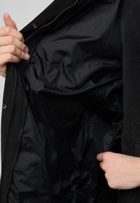 Interno della giacca nera che mostra un tessuto liscio e lucido con una cerniera aperta. Una mano tiene il tirante della cerniera. Presenta un materiale morbido, simile al pile, su un lato.