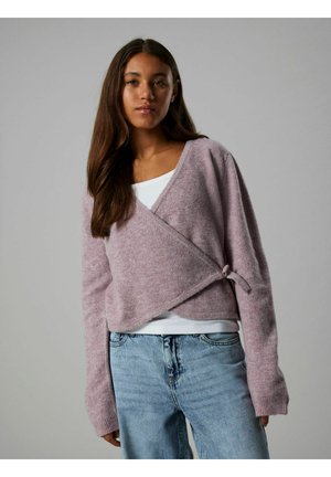 Junge Frau, die einen weichen lila Wickelsweater über einem weißen Oberteil und hellblauen Jeans trägt, steht vor einem schlichten grauen Hintergrund.