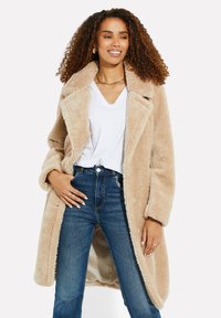 Manteau en fausse fourrure beige avec un large col, fermeture à boutons et poches latérales, associé à un jean bleu taille haute et un tee-shirt blanc à col en V.