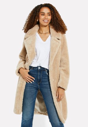 Tan nepbontjas met een brede kraag, knoopsluiting en zijzakken, gecombineerd met hooglopende blauwe jeans en een witte V-hals T-shirt.
