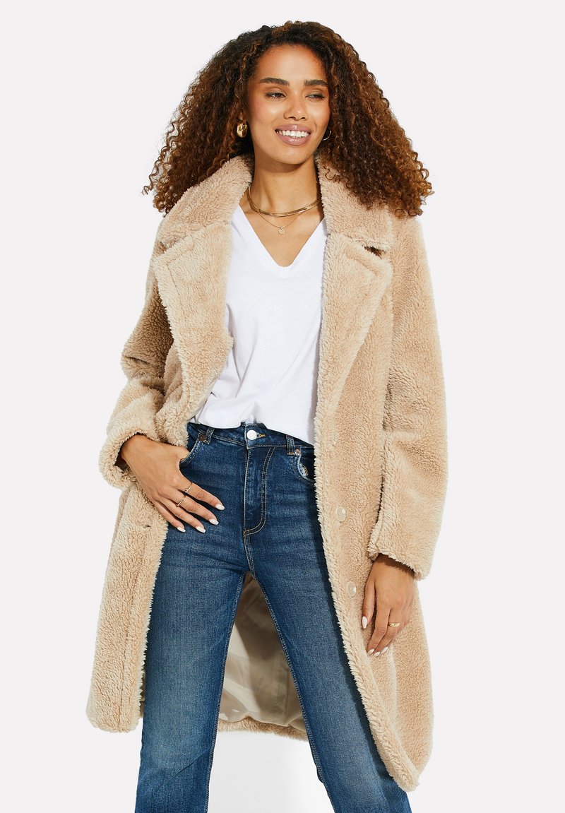 Manteau en fausse fourrure beige avec un large col, fermeture à boutons et poches latérales, associé à un jean bleu taille haute et un tee-shirt blanc à col en V.