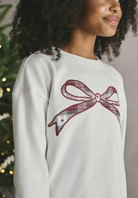 Krämfärgad sweatshirt med ribbad struktur, med en röd och rutmönstrad rosett i mitten. Bakgrunden har gröna lövverk med ljus.