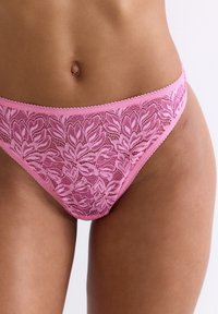 Roze kanten string met bloemmotief, zachte textuur en golvende randen, die de ingewikkelde details en een delicate afwerking benadrukt.