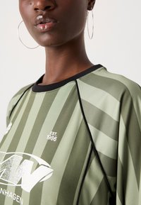 Grün gestreiftes Sportshirt mit schwarzem Halsausschnitt, versehen mit dem Aufdruck "EST. 1986". Der Stoff hat eine glatte Textur und ein sportliches Design.