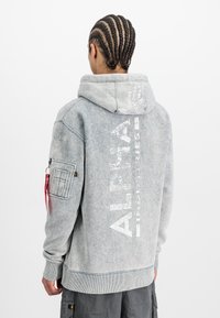 Grau Fleece-Hoodie mit einem abgenutzten Finish, featuring "ALPHA" Logo in Weiß, Reißverschlusstasche am linken Ärmel und geripptem Saum.