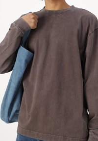 Hnědá košile s dlouhým rukávem vyrobená z měkké tkaniny, s kulatým výstřihem a volným střihem, nošená s modrou džínovou taškou.