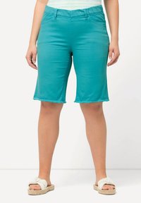 Shorts turchesi realizzati in tessuto elastico con orlo sfilacciato. Fascia elasticizzata in vita e tasche sono visibili. Indossati con sandali bianchi.