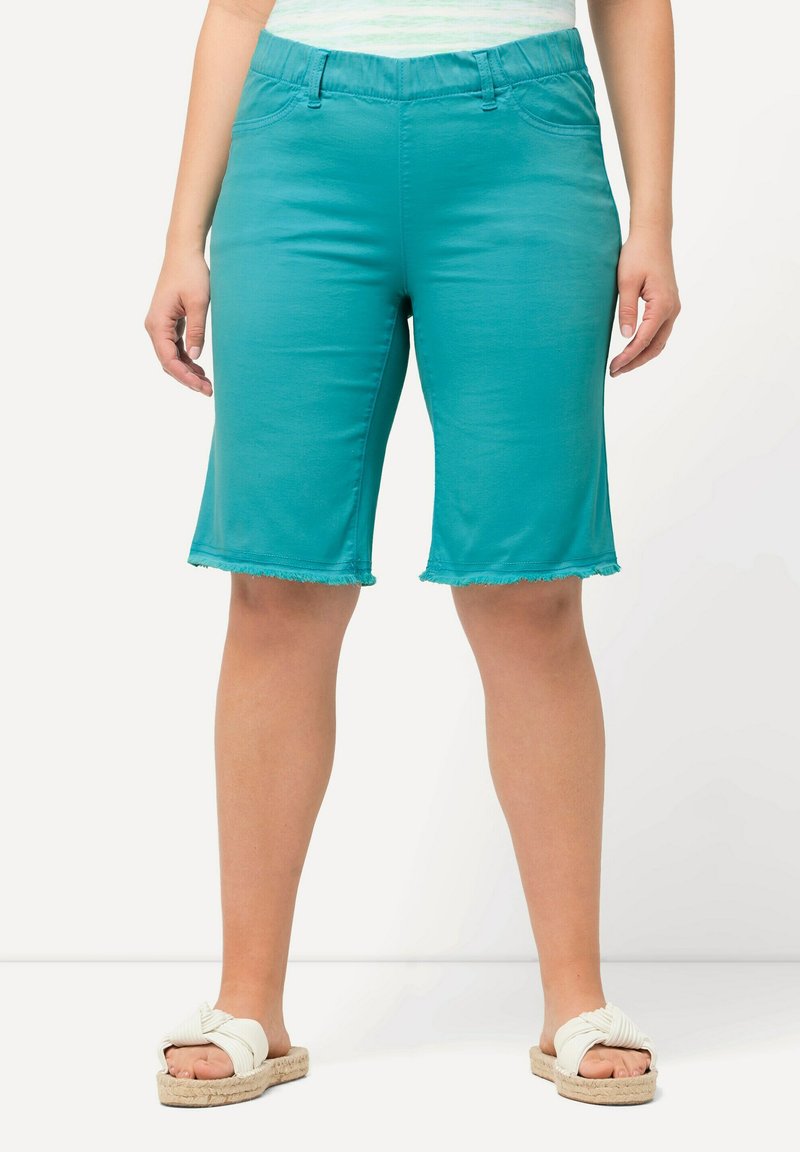Shorts turchesi realizzati in tessuto elastico con orlo sfilacciato. Fascia elasticizzata in vita e tasche sono visibili. Indossati con sandali bianchi.