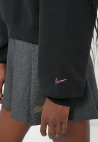 Melns džemperis ar rozā Nike logo uz piedurknes, kombinēts ar pelēku plisētu svārku ar teksturētu apdari.