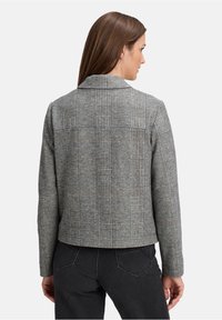 Kobieta z brązowymi włosami ubrana w szary w kratkę żakiet i czarne dżinsy, odwrócona plecami na jednolitym białym tle.