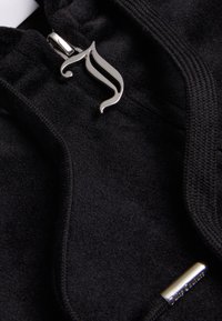 Felpa con zip nera caratterizzata da una cerniera argentata con un design stilizzato, realizzata in tessuto morbido con una consistenza liscia. Decorata con stringhe di regolazione coordinate.