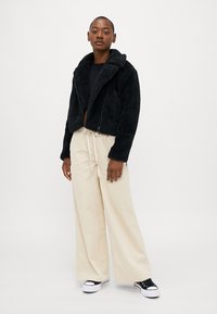 Jaqueta cropped de pêlo sintetico preto com fecho de zipper e colarinho, combinada com calças largas de corduroy bege e ténis pretos.