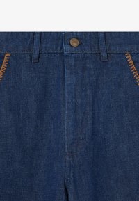 Pantalons en denim bleu foncé avec fermeture à boutons, tissu texturé et surpiqûres orange contrastantes le long des poches et des coutures.