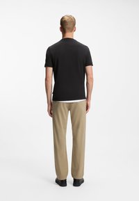 Άνδρας που στέκεται με την πλάτη προς την κάμερα, φοράει μαύρο μπλουζάκι, μπεζ παντελόνι και μαύρα παπούτσια, πάνω σε λευκό φόντο.