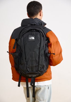 Personne portant un sac à dos noir North Face Hot Shot et une doudoune orange et noire, de dos sur un fond uni.